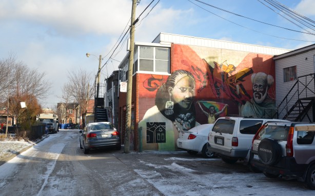 laneway in winter, some snow and ice on the ground.  Three cars parked.  A mural on the side of a two storey building.  Reddish rust coloured background with two faces, one woman and one man. 