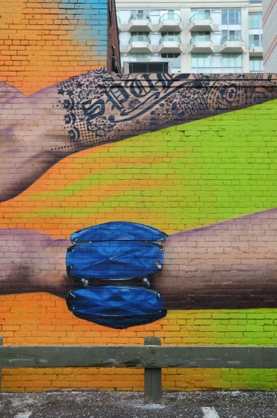 Part of a mural showing wrists of two arms. The upper arm is tattooed in black ink and the tattoo includes the word SPUD.. The bottom wrist is wearing a large blue bracelet. The background is bright green and orange.