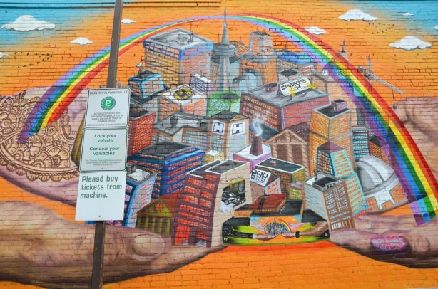 Center part of mural showing a city in 3D under a rainbow, on an orange background, and being held up by four large hands.