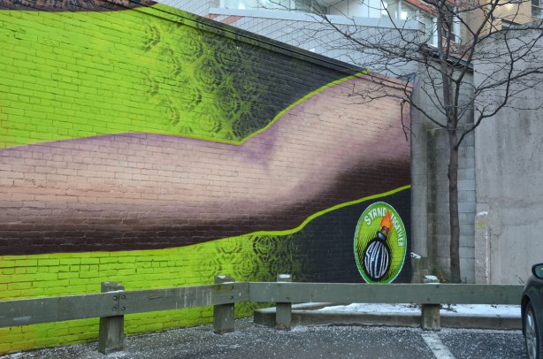 Far right side of mural, behind a low fence, bright green background with a long arm reaching across the photo. A small tree stands in the corner of the parking lot, on the far right of this picture. An apartment building is behind. Spud bomb logo in the bottom right,