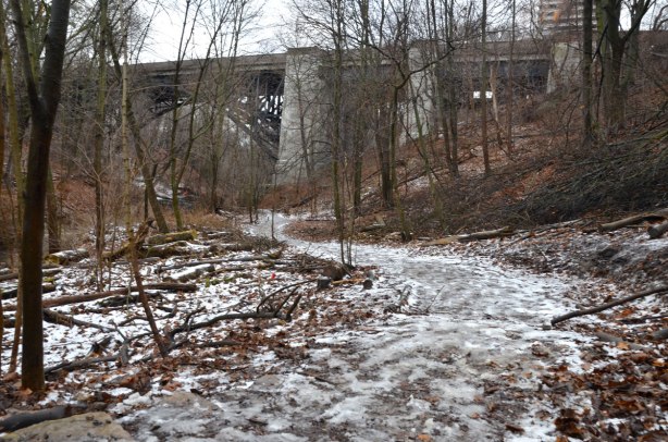 a view of the bridge from a path in the ravine from a short distance away.  It is winter so there is some snow and ice on the path and the trees have no leaves.  