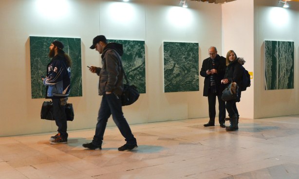 Four people, two are standing together while one texts, and two others are walking past. Four large square paintings (or photos?) are behind them on a white wall.