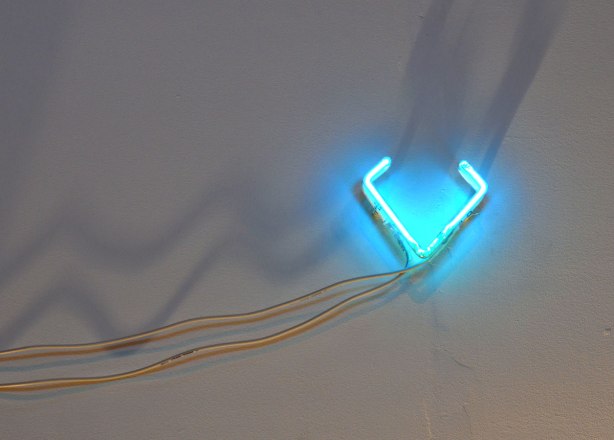 Neon art piece on a wall. It is shaped like a V, it's turquoise blue in colour and it is a bit bigger than the size of a hand. It is mounted on a wall with the electrical wires visible. It is shadow.