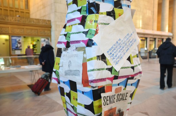 part of a piece of art that is a pillar like structure cover with bits of colourful paper. There is also a child's drawing in pencil on an 8 by 11 piece of paper. Also a sign in blue letters that says . In the background there are a couple of people walking past.