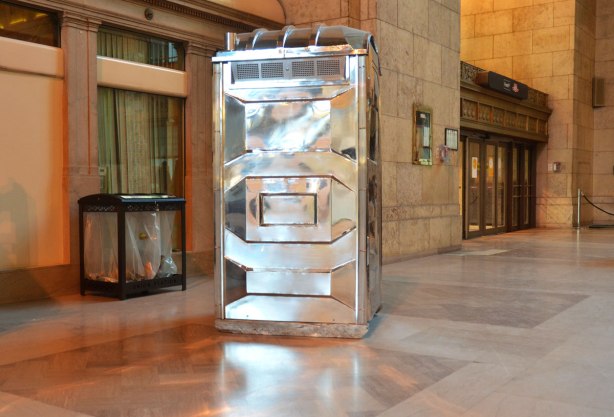 An art piece, a shiny silver coloured porta potty stands in the middle of the floor. A trash container (real) is behind it.