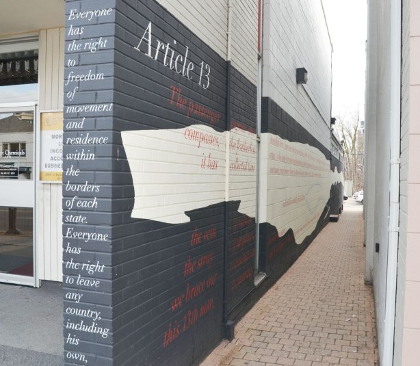 A narrow lane, or pedestrian walkway, between two buildings.  On the left hand side wall there is a mural with a black and white background, and red letters.  The words are a poem about freedom of movement as stated in article 13 of the Declaration of Human Rights. 