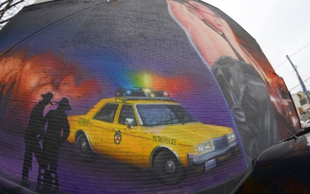part of a large mural showing a yellow Toronto police car from the 1980's, a couple of policemen and a crowd of men standing just back of the police car