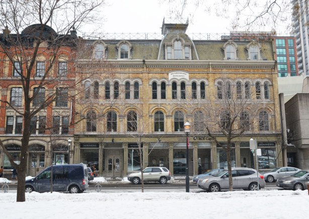 Four storey tall brick building with arched windows and mansard roof, yellowish brick.  White and black stone sign built into the building at the level of the third floor that says 'Beardmore Building'.