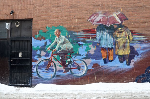 Part of a mural on an alley wall.  There are two parts of the mural visible in this photograph.   An older man is riding a bicycle in one part and twp people are sharing an umrella and walking away from the viewer in the other part