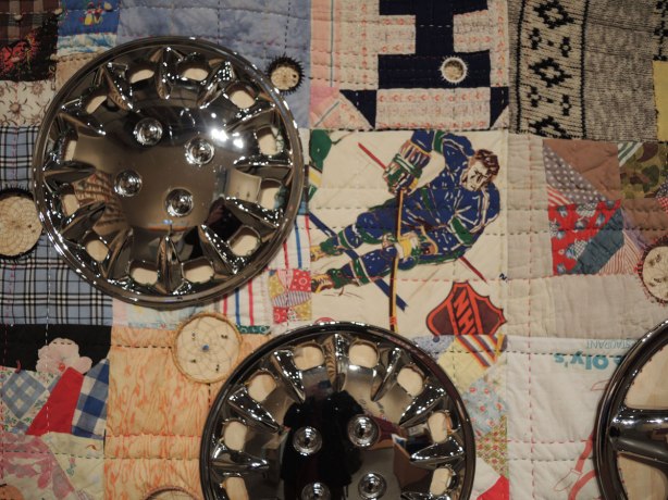 close up of part of a quilt.  One of the squares is made from fabric with a Toronto Maple Leafs hockey player on it.  Old chrome hubcaps have been added to the quilt. 