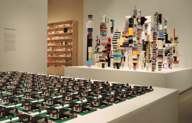 A portion of three Douglas Coupland creations are visible.  In the foreground are numerous lego houses complete with yards and garages all laid out in a grid.  In the background are wood shelves with household articles from the past.  In the middle are lego towers in bright stripes and interesting shapes. 