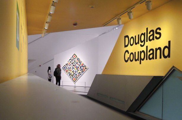 entrance to an exhibit at the Royal Ontario Museum featuring a large yellow wall with the name Douglas Coupland in large black letters.  In the distance are two women standing in front of a painting that is hung on point. 