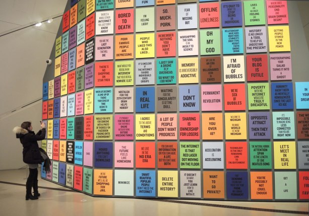 a large wall is covered with coloured rectangles and in each rectangle is an expression that has become common.  For example, get a life, oh my god, delete entire history?, etc.  A woman is standing to the left of the wall, taking a picture of it with her cellphone. 