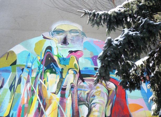 A graffiti face high on a grey wall, with a pine tree branch  partially obscuring it