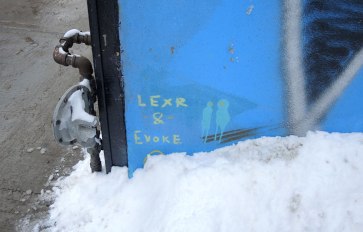 In the corner of a piece of graffiti with a blue background is a signature of the artists, in blovk letters, LEXR & EVOKE