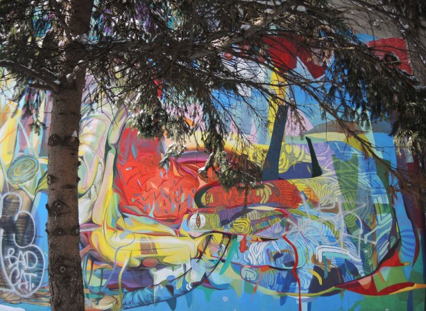 Close up a piece of street art featuring brightly coloured fingers, with eyes on the fingernails.  Partially hidden behind pine trees.  Snow on the ground. 