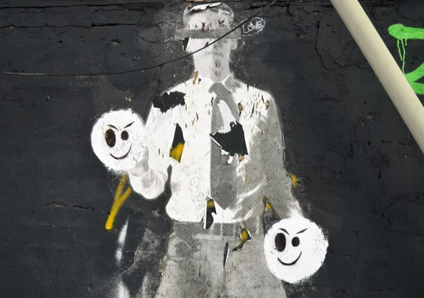 black and white paper graffiti of a man in grey pants, white shirt, and tie.  He has two black and white faces instead of hands.  Someone has scrawled words on the hands. 