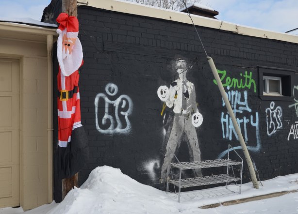 A deflated inflatable Santa Claus is hanging from a hydro pole in an alley beside a garage door with a graffiti man on it. 