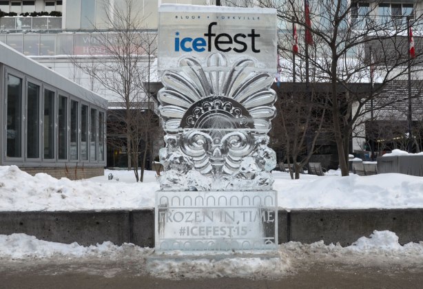 Ice sculpture beside a sidewalk.  The top part has the words Bloor Yorkville icefest and the bottom part has the words Frozen in time, icefest 15.   The middle section looks like a large face made of Egyptian motifs. 