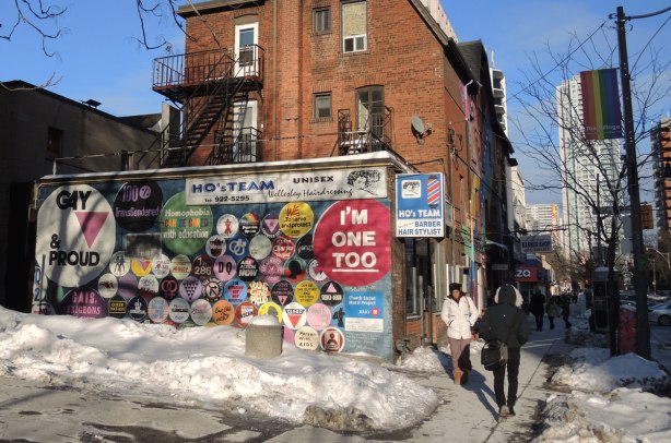 street scene in winter, looking along the sidewalk with a couple of people on it.  On the left is a three storey red brick building in the background.  In the foreground is a shorter building (seen from the side) covered with a mural depicting buttons with slogans and sayings on them. 
