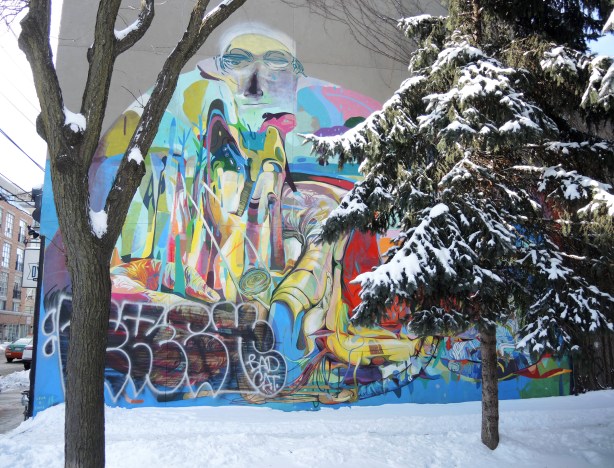 Graffiti painting of a man high up on a grey wall, partially hidden behind two trees.  Unfortunately someone has painted a black tag over the lower left corner of the painting