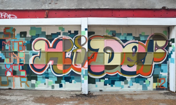 Graffiti painting on a garage door of the words "Hi Def" in rounded fat letters with colours in pinks greens and black squares. 