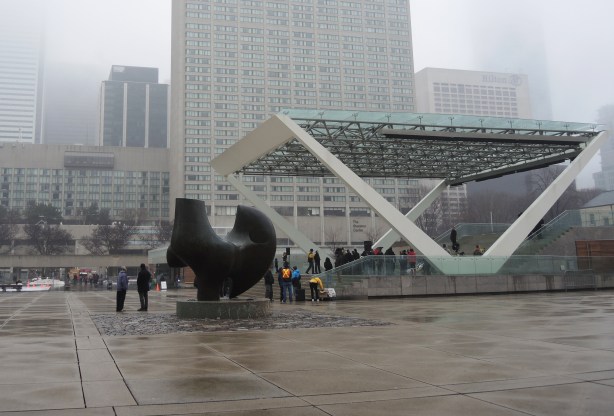 Nathan Phillips square on a foggy wet day.  Greyness.  There are a few people under the stage overhang but otherwise the square is empty