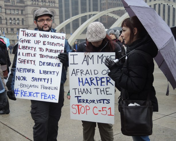 A group of 3 people.  A woman on the right is holding a purplish grey umbrella.  The other 2 people are holding protest signs. 