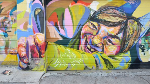 multicoloured street art on a garage door of a boy reaching forward, his hand is foreshortened and is clasest to the viewer. 