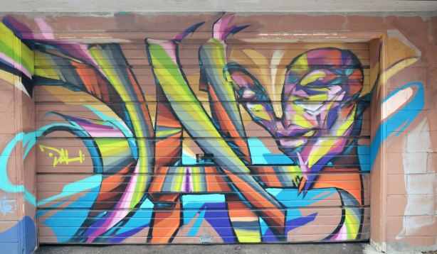 Graffiti on a garage door.  A face is in the top right corner and she is looking down towards the ground.  Stylized. 