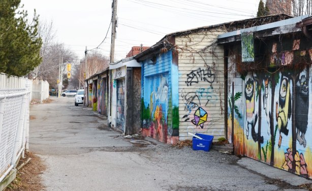 north end of an alley with garage doors on the right, white chain link fence on the left.   The garage doors are covered with street art and graffiti. 