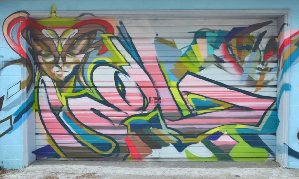 Street art piece of a tag in the middle and a face on either top corner. 