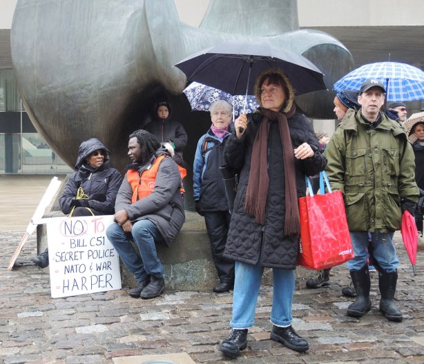 A group of people around Henry Moore's sculpture "the Archer" in Nathan Phillips Square.  They are there for the anti Bill C51 protest.  A couple are sitting and talking to each other.  Others are standing under umbrellas. 