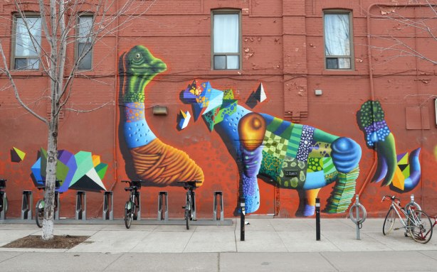 birdo street art on a rust coloured brick wall. Two animal like creatures in greens, blues and rusts. They are larger than life size, taking up most of the side of the building, up to the second storey level.
