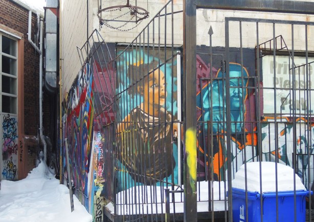 Two graffiti faces on a wall, one woman and one man. Metal bars surrounding a small parking spot behind the building partially obscure the wall.