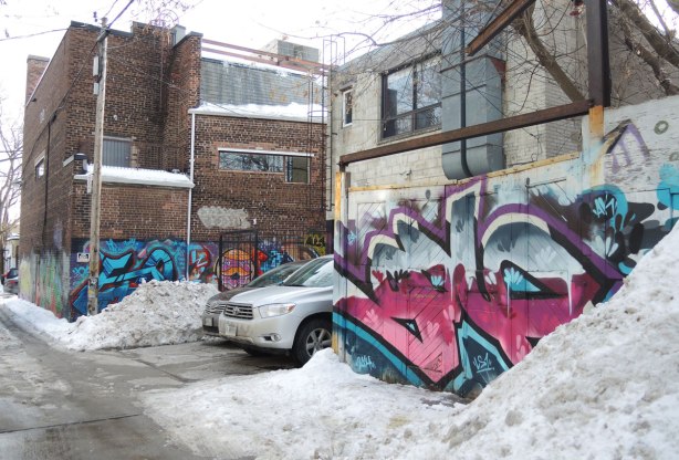 backs of buildings, cars parked behind the buildings, piles of snow on the side of the alley, graffiti on the walls and garage doors. 