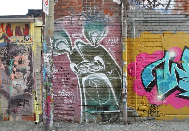 large greenish brown poser bunny in Graffiti Alley on pinkish purple background.   Bight yellow, pink and blue tag to the right and a door covered with small tags to the left 