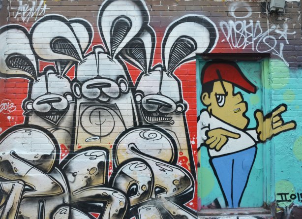 three black and white poser bunnies on red background with black and white tag in front  in Graffiti Alley.  They are beside a doorway on which a man has been painted - red hair, white T-shirt and blue pants.  Stylized, shrugging his shoulders and holdin his hands at waist level. 