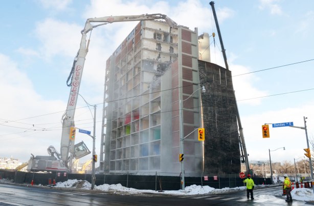 Two very large cranes are being used to demolish a large apartment building on the corner of Dundas and Sumach streets.  Some men in bright yellow vests are directing traffic as some of the debris is falling towards Sumach street. 