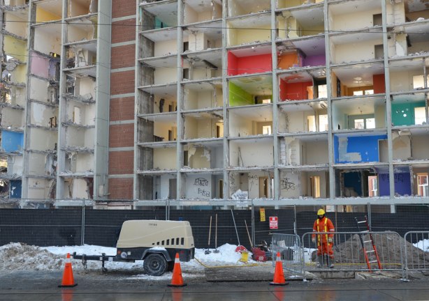 A workman dressed in orange coveralls works on the street in front of a building being demolished