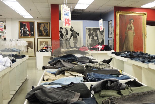 Jeans for sale, on tables in Honest Eds store.  Large black and white posters on the wall along with a colour full length portrait of a woman in a long dress. 