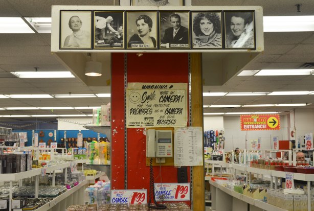 kitchen wares for sale laid out on white table like shelves.  Large pillar in the middle of the store with a sign warning you that you are on camera.  Seven pictures of movie stars adorn the pillar.  Lots of merchandise for sale in the background. 