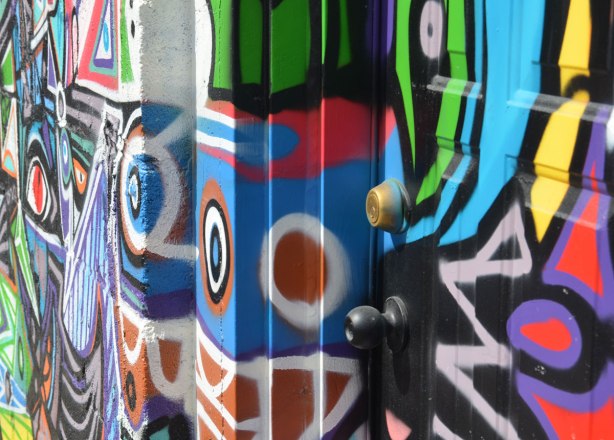 Close up, and a side angle shot, of colourful graffiti on a door, door frame and wall. Bright colours, geometric shapes such as white circle around brown, pink zig zags.