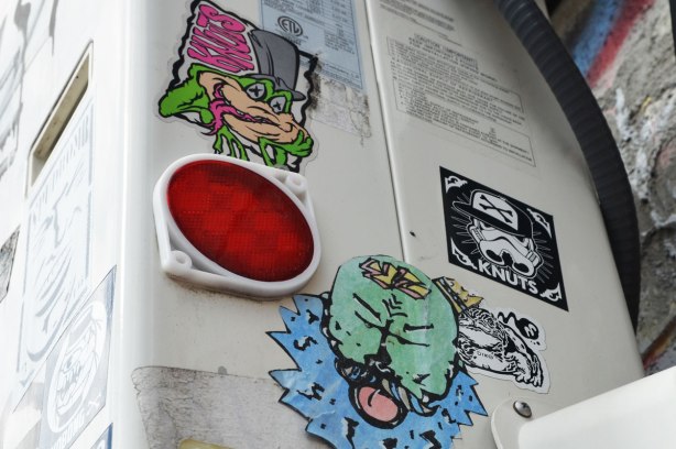 Some stickers or slaps on the side of an air conditioner. One is of a frog head with its tongue out and eyes closed. The other is of a green man's face, also with his tongue out.