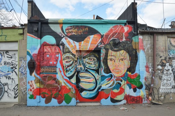 A wall with a door in the middle of it. There is a street art piece of three heads. Two very stylized men and a more realistic looking woman. Bright colours.