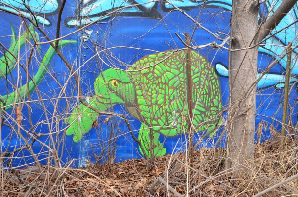 Part of a mural showing a large green turtle swimming in a creek.  A tree with no leaves is in front of the mural 