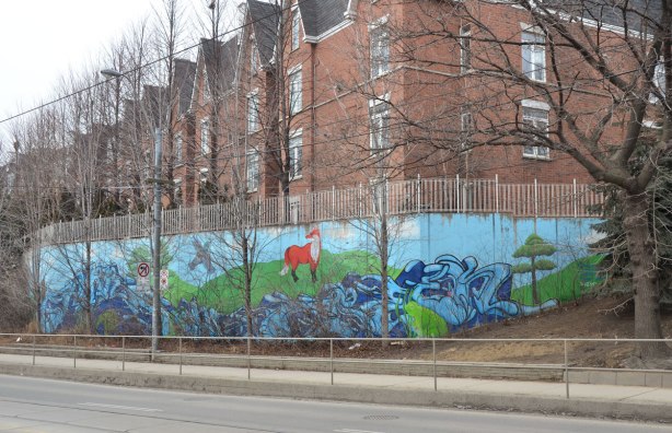 A mural is painted on a retaining wall at the bottom of a small hill that separates some row houses and the street.  Many small trees are in front of the mural but it is winter time so there are no leaves on the trees.  The mural is a stylized creek that flows through hills with some animals standing beside it - fox, frog, turtle and beaver. 