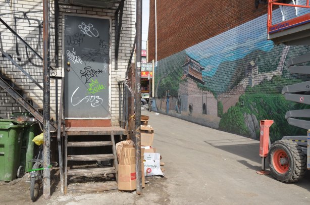part large mural of the great wall of China in an alley, the street end of the alley with part of the mural, the back door of a shop on the other side of the alley is also in view