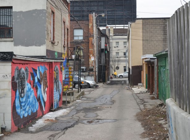 looking west along an alley in late winter.  Street art on the walls on the left, garage doors on the right.  Buildings on Roncesvalles can be seen at the end of the alley which is about a block long. 