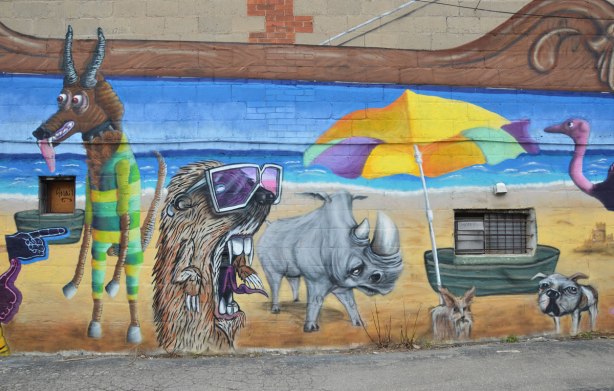Destination Mammals Cabana mural, hairy creature with mouth wide open and wearing suglasses and a rhino by a beach umbrella, street art, graffiti, 